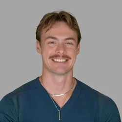 Austin Smith smiling, with light brown hair and a mustache, wearing teal scrubs and a necklace against a gray background.