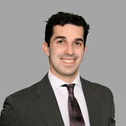 Ryan Neydavood smiling, with short curly dark hair, wearing a gray suit, white shirt, and dark tie against a gray background.