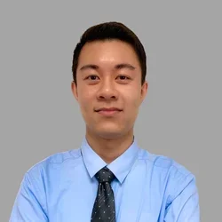 Ryan Tran with short dark hair and glasses, wearing a light blue shirt and dark tie against a gray background.
