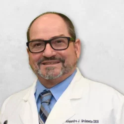 Dr. Urdaneta is wearing a white doctor's coat over a light blue shirt with a tie. He is also wearing glasses.
