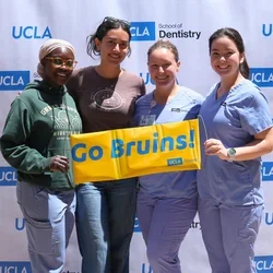 Four smiling dental students, holding a yellow sign reading "Go Bruins!"
