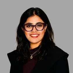 Ahana Goswami smiling, with long wavy dark hair and glasses, wearing a black jacket over a dark top against a gray background.