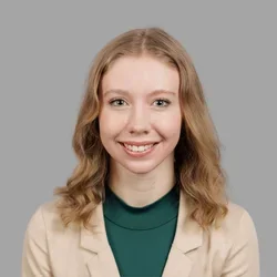 Christina Lindberg smiling, with shoulder-length wavy blonde hair, wearing a beige blazer over a green top against a gray background.