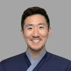 Paul Chung smiling, with short dark hair and light facial hair, wearing a blue coat against a gray background.