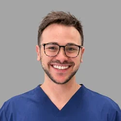 Ariel Oren smiling, with short brown hair, glasses, and a trimmed beard, wearing blue scrubs against a gray background.