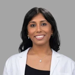 Shalini Sharma smiling, with shoulder-length dark hair, wearing a white coat over a dark top and a necklace against a gray background.