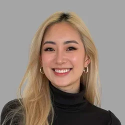 Sammy Gong smiling, with long blonde hair, wearing a black turtleneck and hoop earrings against a gray background.