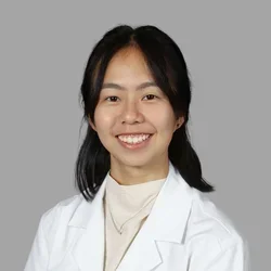Yen Tung Lieu smiling, with dark hair pulled back, wearing a white coat over a light top against a gray background.