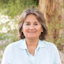 Dr. Diana Messadi wearing a light blue button-up shirt. She also has a gold necklace with a geometric pendant and small gold stud earrings.
