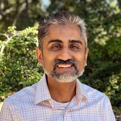 Dr. Mohammed Husain, smiling in a plaid shirt with some green bushes in the background