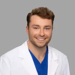 Adam Fuller smiling, with short brown hair and light facial hair, wearing a white coat over blue scrubs against a gray background.