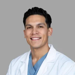 Ali Hernandez smiling, with short dark hair, wearing a white coat over blue scrubs against a gray background.