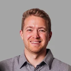 Arick Christopher smiling, with short reddish-brown hair, wearing a gray collared shirt against a gray background.