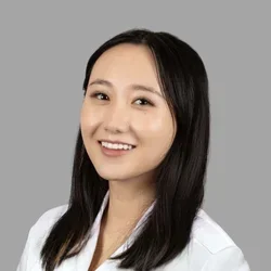 Chelsey Chen smiling, with straight shoulder-length dark hair, wearing a white coat against a gray background.
