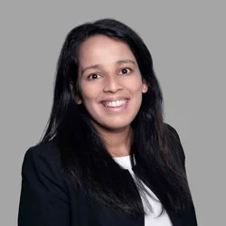 Shveta Duggal smiling, with long dark hair, wearing a dark blazer over a light top against a gray background.