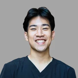 Brendan Co smiling, with short dark hair, wearing dark scrubs against a gray background.
