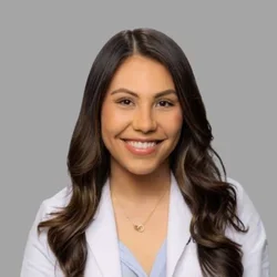 Sarah El Rahmany smiling, with long wavy dark hair, wearing a white coat over a light blue blouse against a gray background.