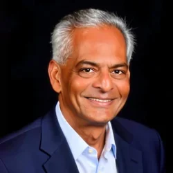 Dr. Shetty is posing in front of a dark background, wearing a dark blue suit and a light blue collared shirt.