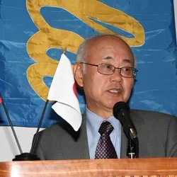 Dr. Henry Takei speaking at a podium, with small USA and Japanese flags next to him and a UCLA banner behind him.