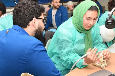 A dental student in blue coat instructing another student in green gown