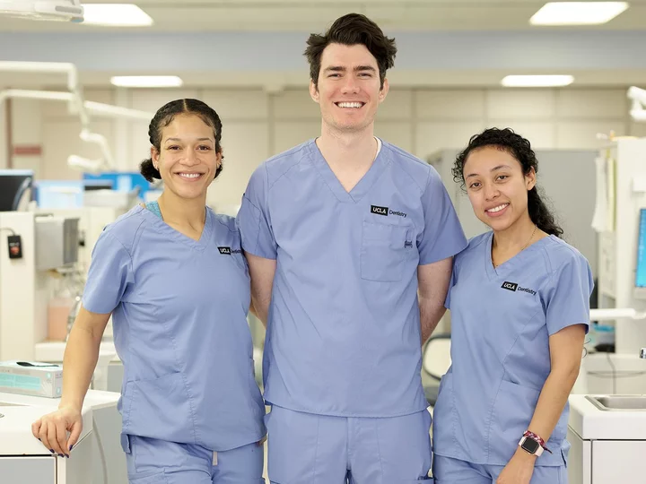 Three smiling UCLA dental students in blue scrubs in a clinic.